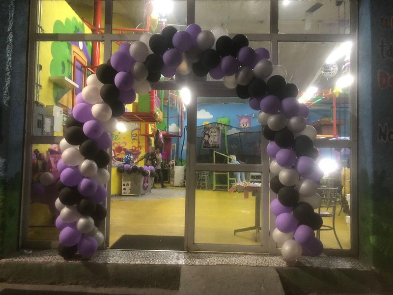 Entrada de Tadeopolis de noche con arco de globos morado, negro y plateado iluminado