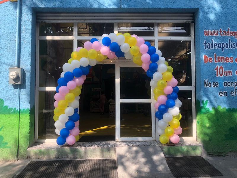 Entrada del salón con arco de globos azul, amarillo, rosa y blanco en la fachada